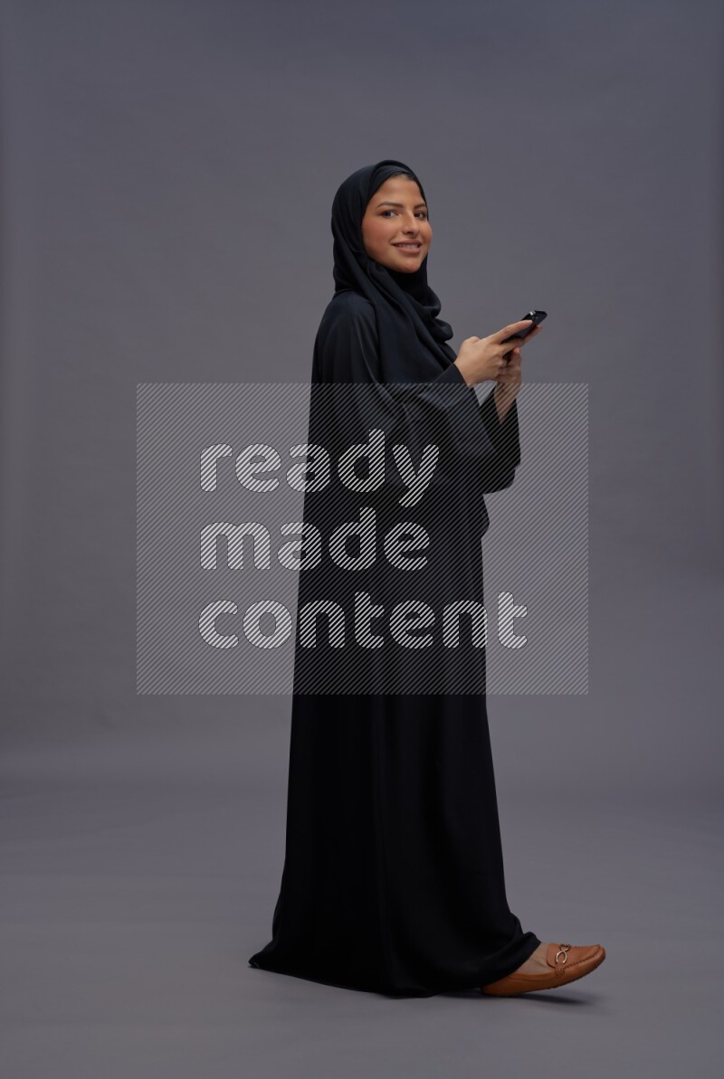 Saudi woman wearing Abaya standing texting on phone on gray background