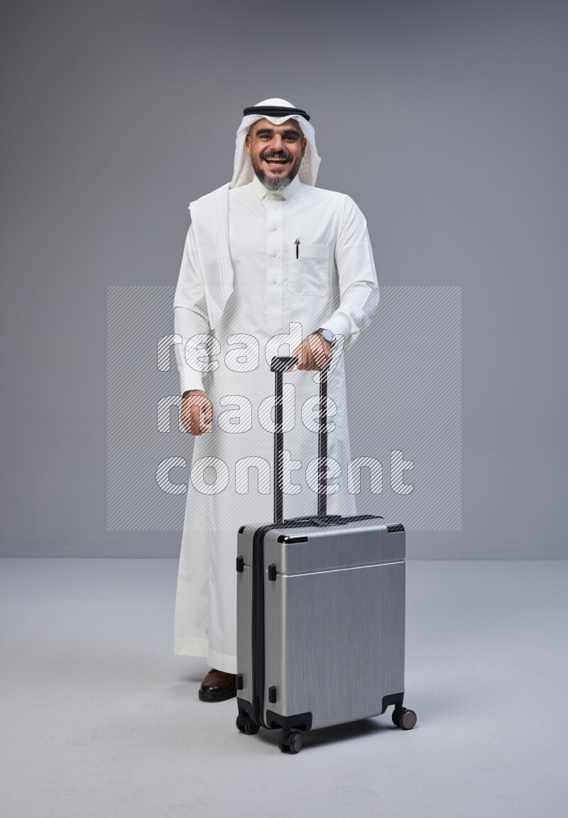 Saudi man wearing Thob and white Shomag standing holding Travel bag on Gray background
