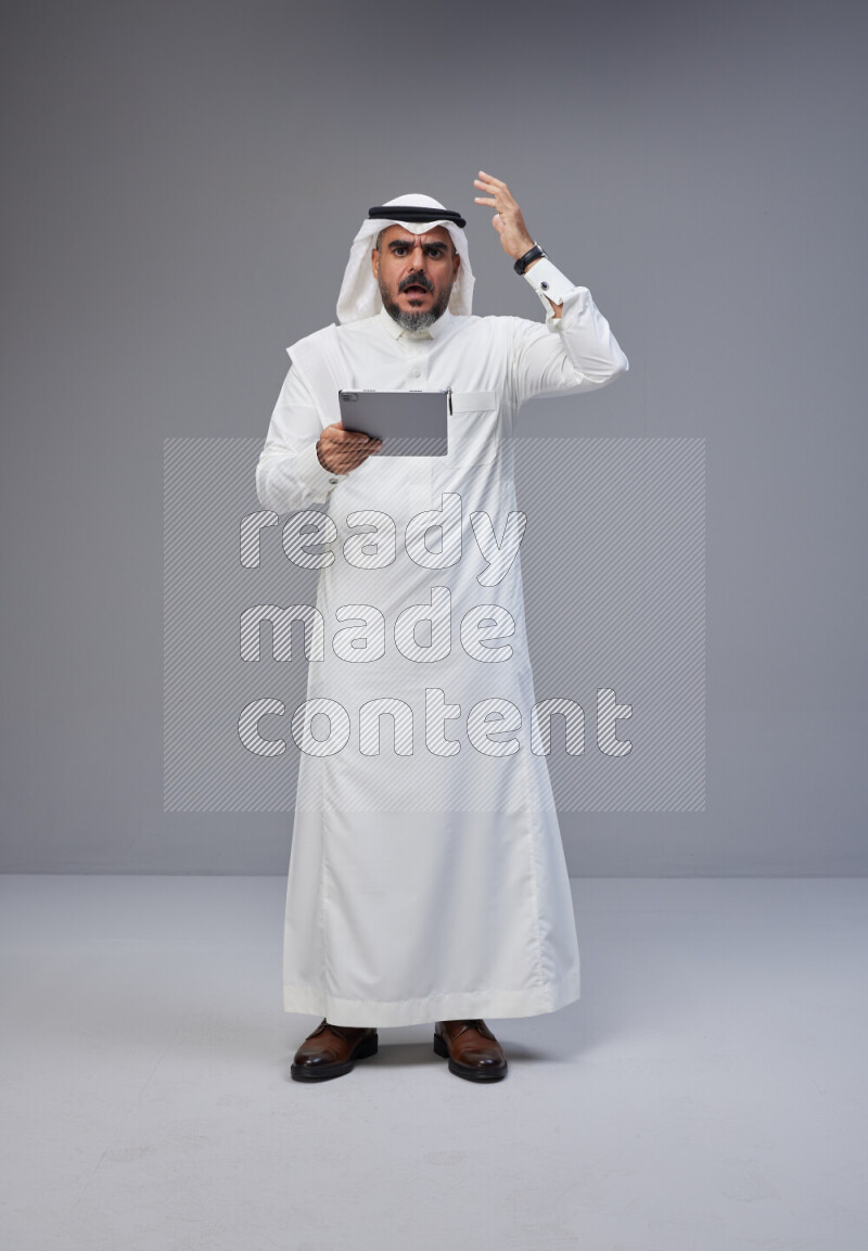 Saudi man Wearing Thob and white Shomag standing working on tablet on Gray background