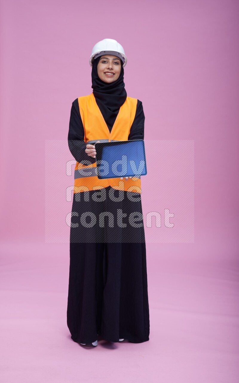 Saudi woman wearing Abaya with engineer vest and helmet standing showing tablet to camera on pink background