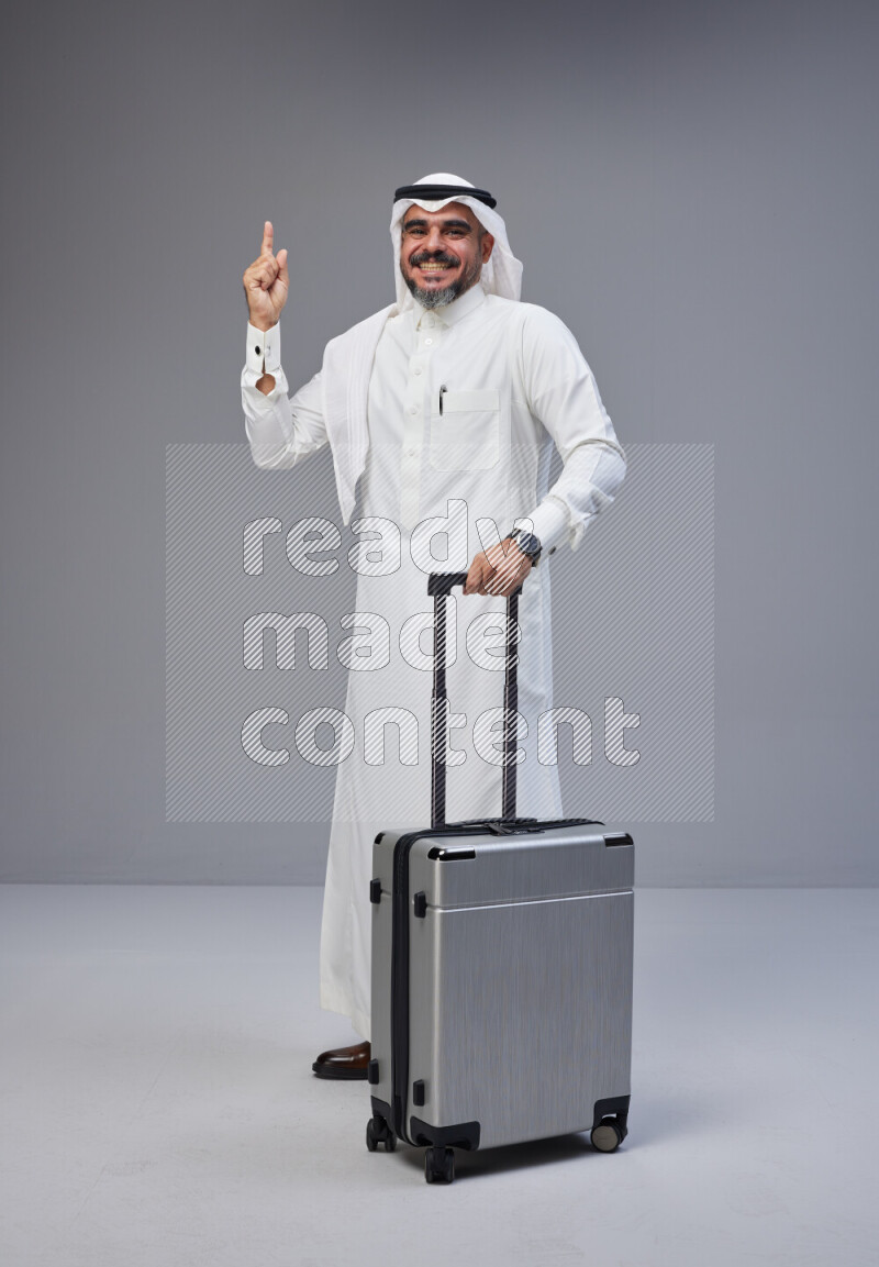 Saudi man wearing Thob and white Shomag standing holding Travel bag on Gray background