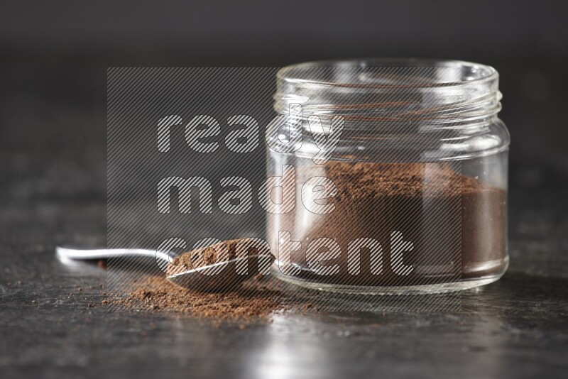 A glass jar full of cloves powder with a metal spoon on a textured black flooring