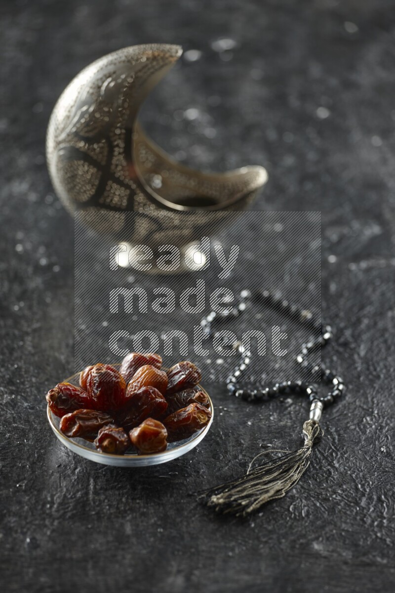 A silver lantern with different drinks, dates, nuts, prayer beads and quran on textured black background