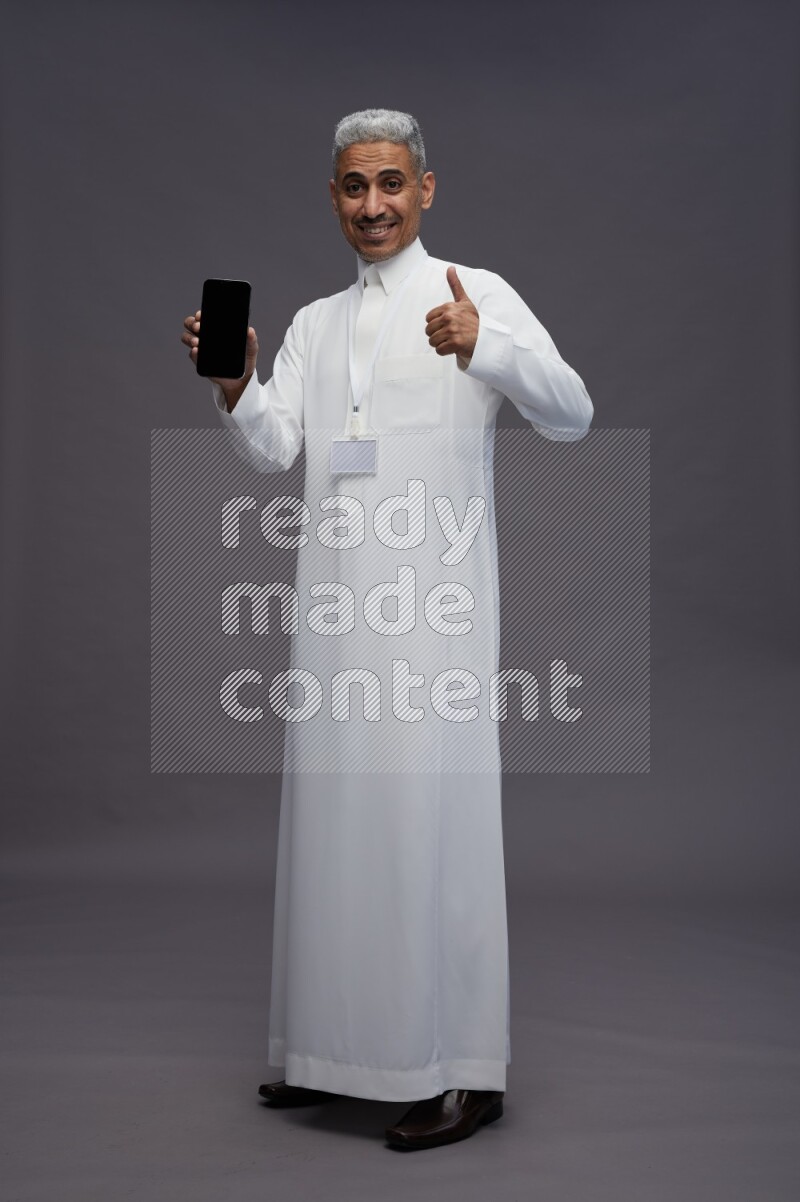 Saudi man wearing thob with neck strap employee badge standing showing phone to camera on gray background