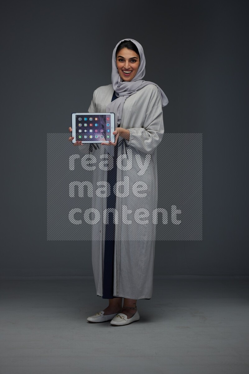 A Saudi woman wearing a light gray Abaya and head scarf standing and showing the phone's screen eye level on a grey background