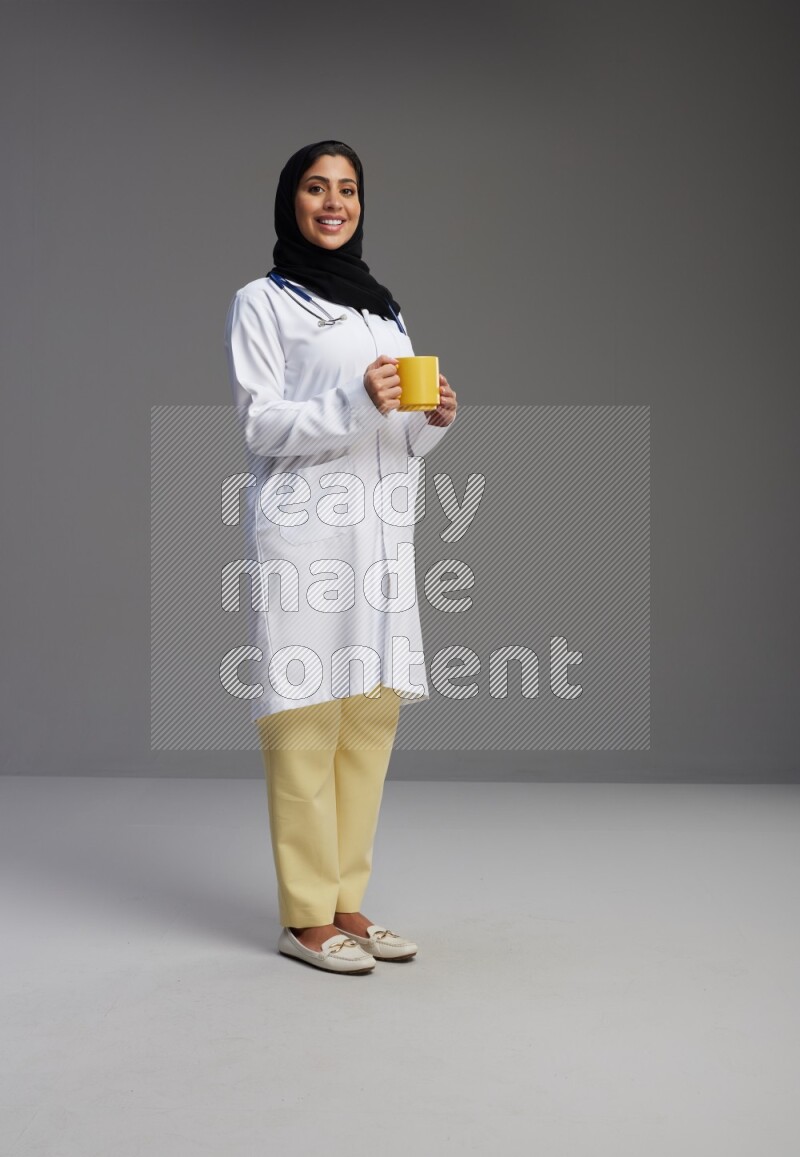 Saudi woman wearing lab coat with stethoscope standing holding mug on Gray background