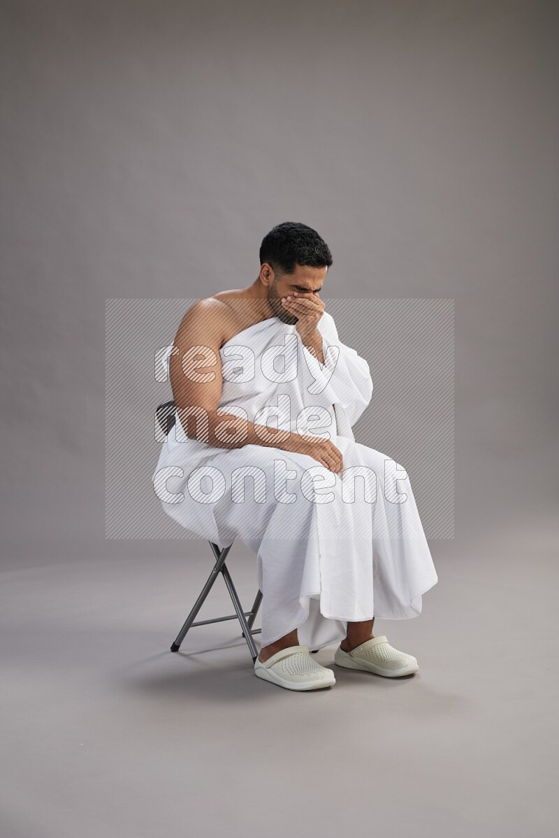 A man wearing Ehram sitting on chair showing how to sneeze on gray background