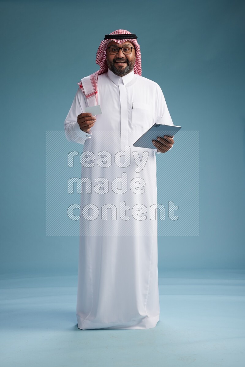 Saudi Man with shimag Standing holding ATM card while working on tablet on blue background
