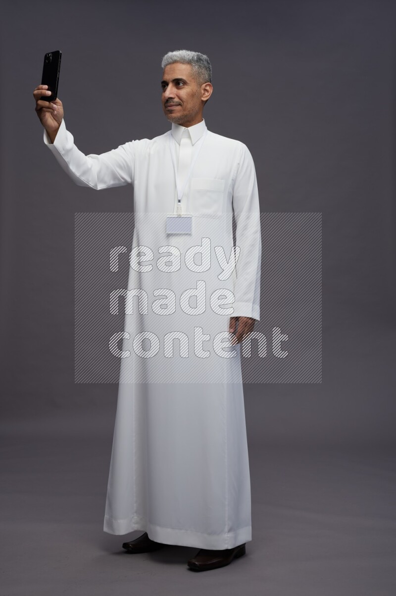 Saudi man wearing thob with neck strap employee badge standing taking selife on gray background