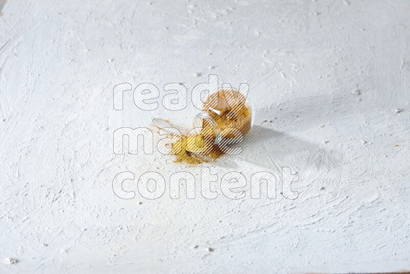 وعاء زجاجي للتوابل مقلوب وملعقة معدنية ممتلئان ببودرة الكركم والبودرة تناثرت منه على خلفية بيضاء