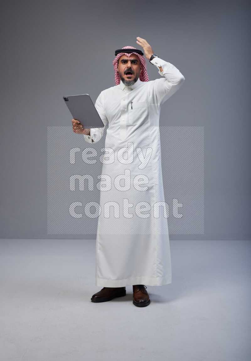 Saudi man Wearing Thob and red Shomag standing working on tablet on Gray background