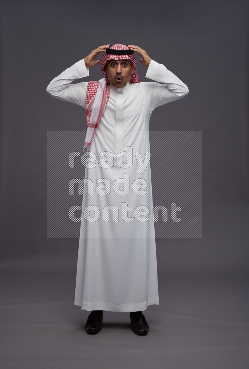 Saudi man wearing thob and shomag standing hands behind head on gray background