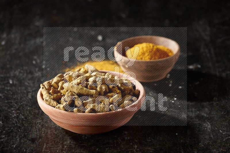 2 wooden bowls, one full of turmeric powder and the other full of dried turmeric whole fingers on a textured black flooring