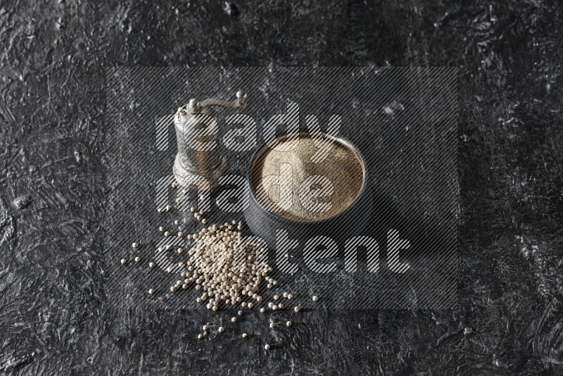 Black pottery bowl full of white pepper powder with pepper beads and metal pepper grinder on textured black flooring