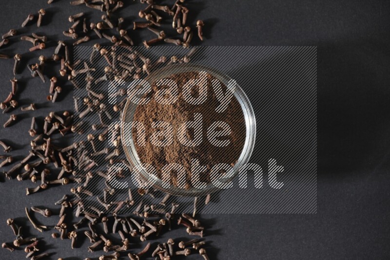 A glass bowl full of cloves powder with cloves grains spread on black flooring