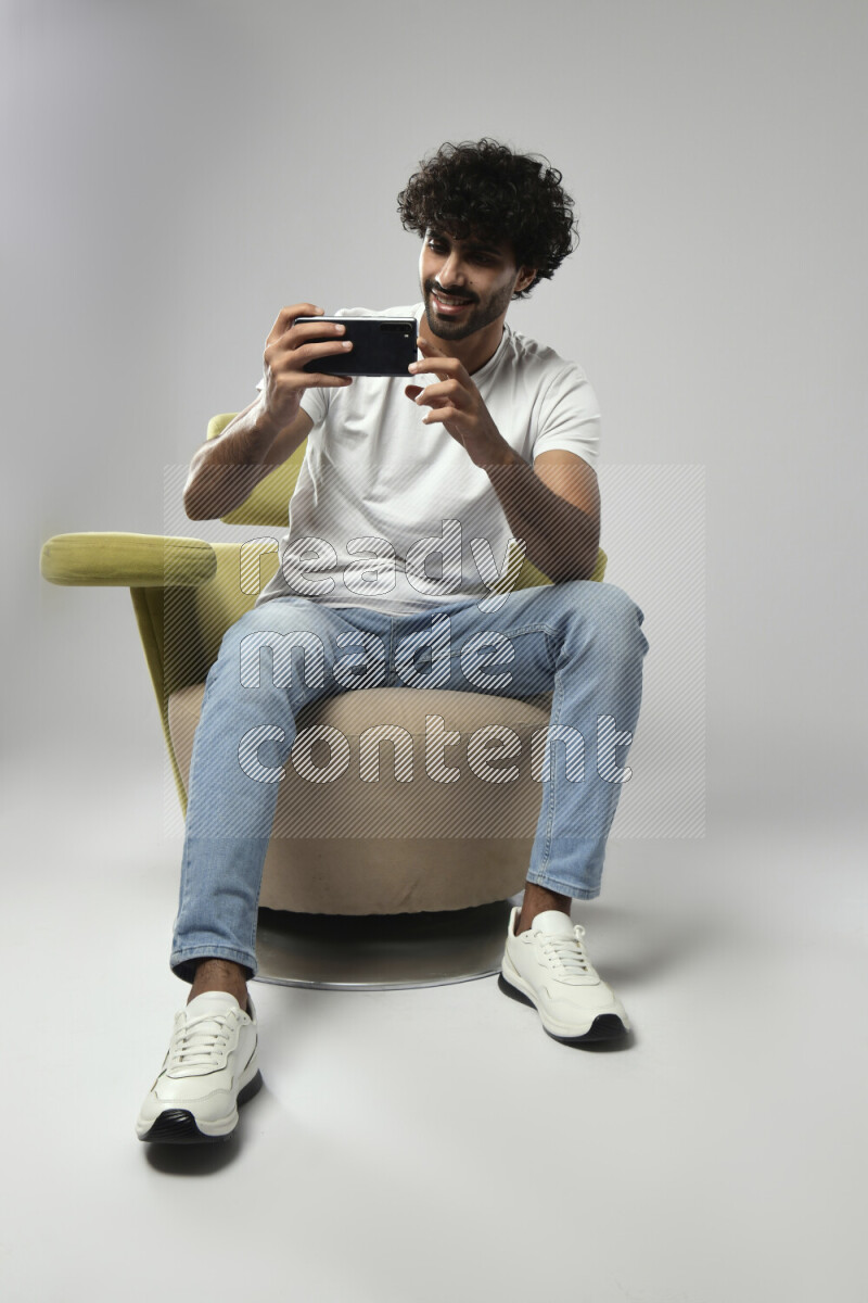 A man wearing casual sitting on a chair shooting with his phone on white background