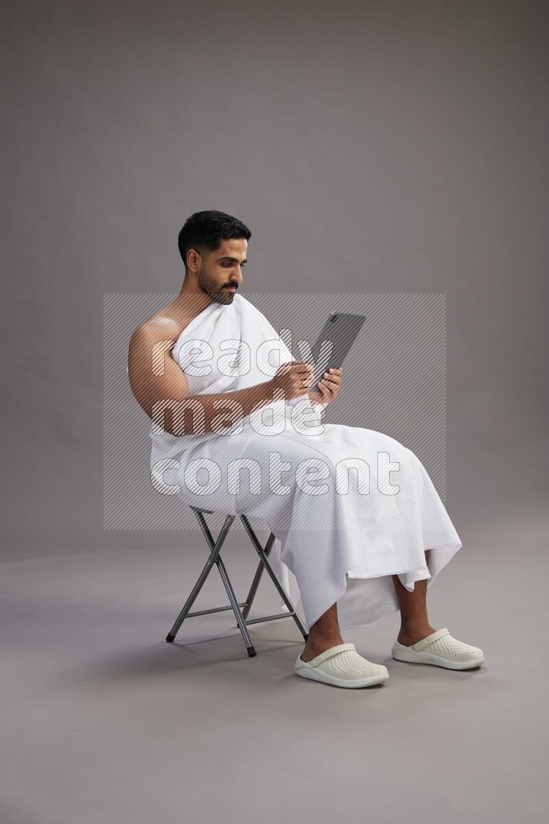 A man wearing Ehram sitting on chair holding a tablet on gray background