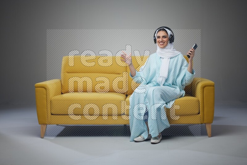 A Saudi woman wearing a light blue Abaya and a white head scarf sitting on a yellow sofa and holding an iPad while wearing headphone eye level on a grey background