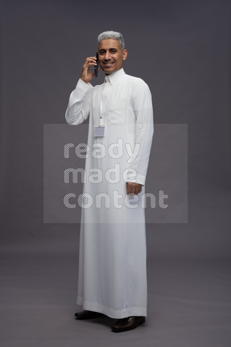 Saudi man wearing thob with neck strap employee badge standing talking on phone on gray background