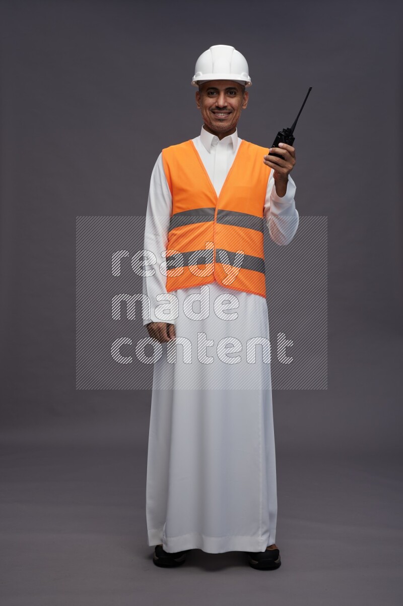 Saudi man wearing thob with engineer vest standing holding walkie-talkie on gray background