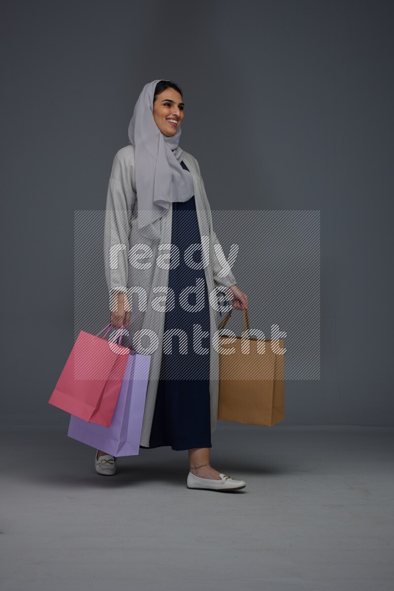 A Saudi woman wearing a light gray Abaya and head scarf standing and holding shopping bags on a grey background