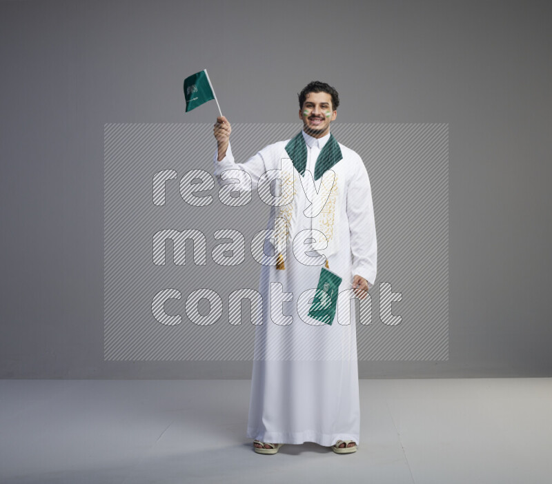 A Saudi man standing wearing thob and saudi flag scarf with face painting holding small Saudi flag on gray background