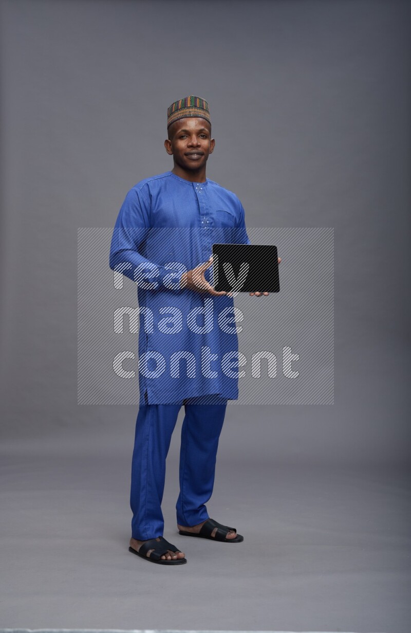 Man wearing Nigerian outfit standing showing tablet to camera on gray background