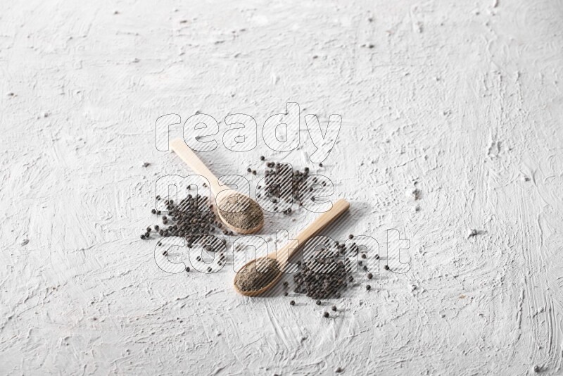 2 wooden spoons full of black pepper powder and black pepper beads spread on textured white background