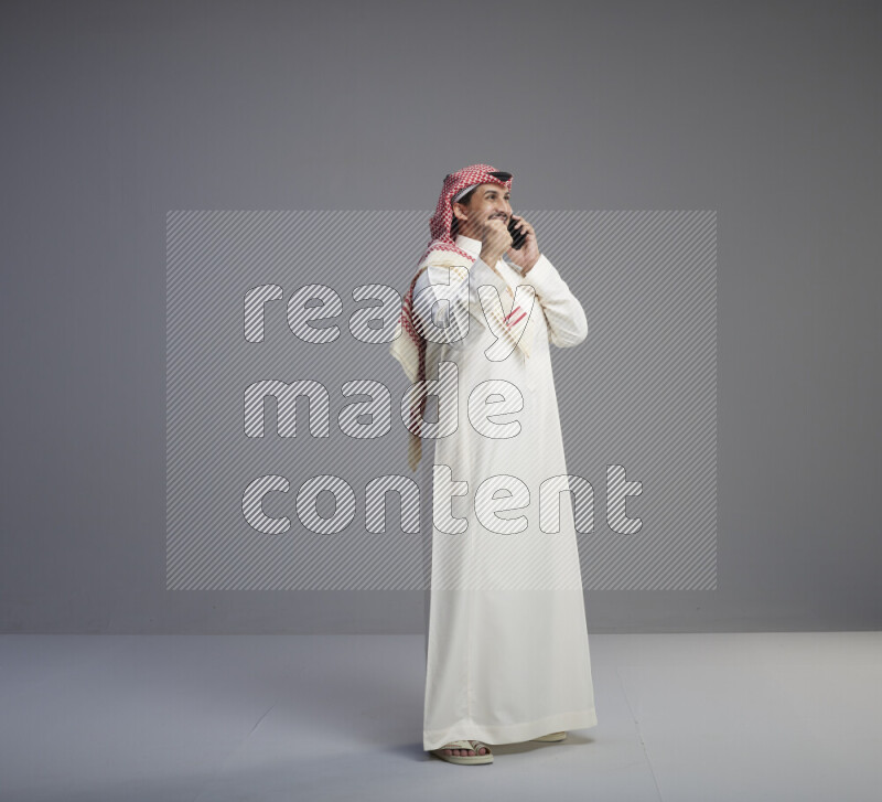 A Saudi man standing wearing thob and red shomag talking on phone on gray background