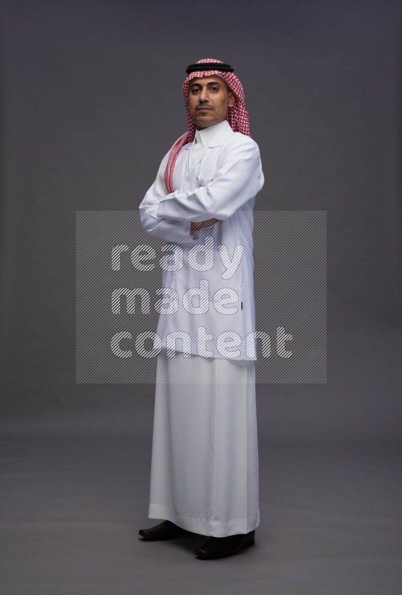 Saudi man wearing thob with lab coat and shomag with pocket employee badge standing with crossed arms on gray background