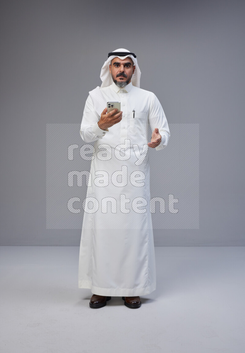 Saudi man Wearing Thob and white Shomag standing texting on phone on Gray background