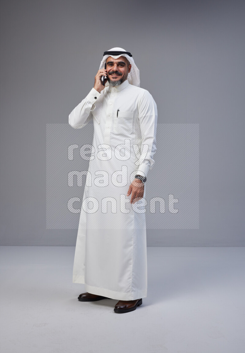 Saudi man Wearing Thob and white Shomag standing talking on phone on Gray background