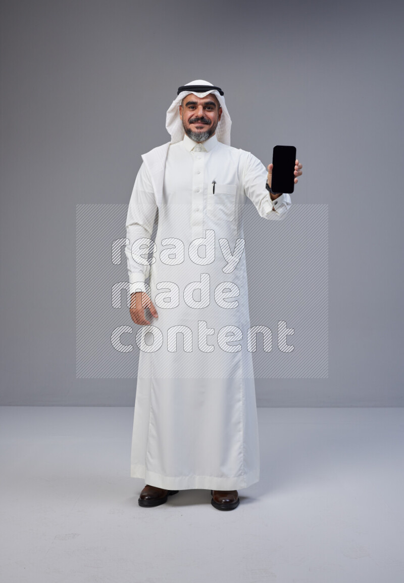 Saudi man Wearing Thob and white Shomag standing showing phone to camera on Gray background