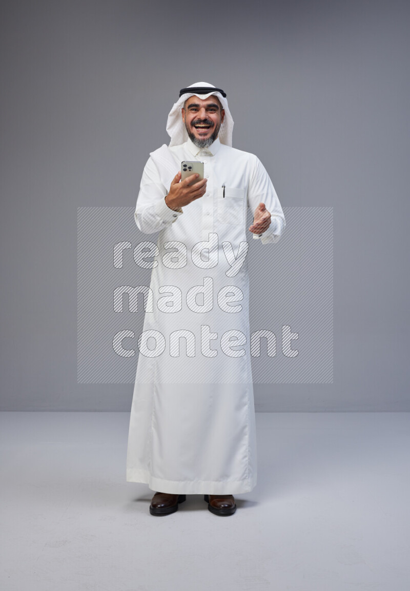 Saudi man Wearing Thob and white Shomag standing texting on phone on Gray background