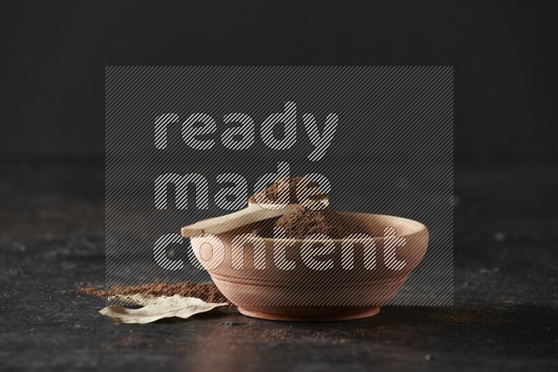 A wooden bowl and a wooden spoon full of cloves powder with laurel leaves on a textured black flooring