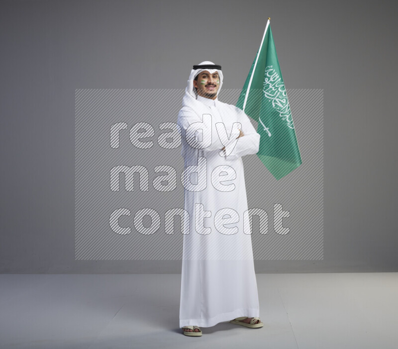 A Saudi man standing wearing thob and white shomag with face painting raising big Saudi flag on gray background