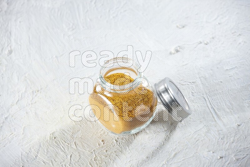 A glass spice jar full of turmeric powder on a textured white flooring