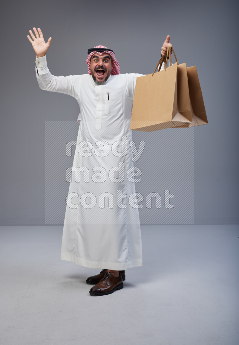 Saudi man Wearing Thob and red Shomag standing holding shopping bag on Gray background