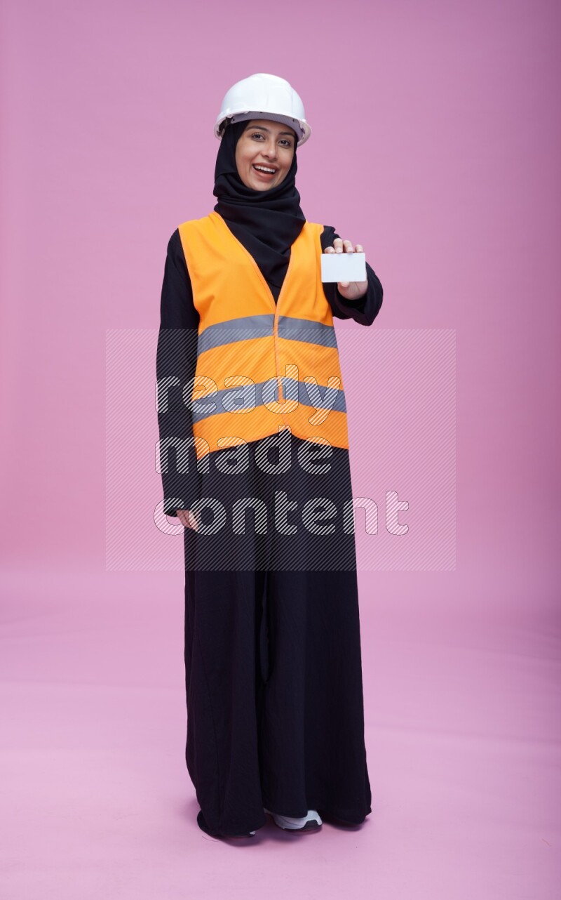 Saudi woman wearing Abaya with engineer vest and helmet standing holding ATM card on pink background