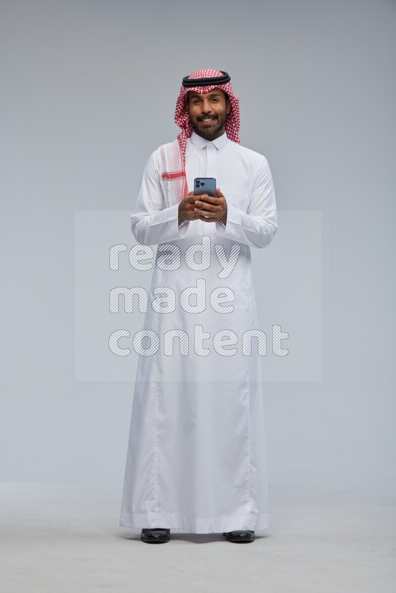 Saudi man Wearing Thob and shomag standing texting on phone on Gray background