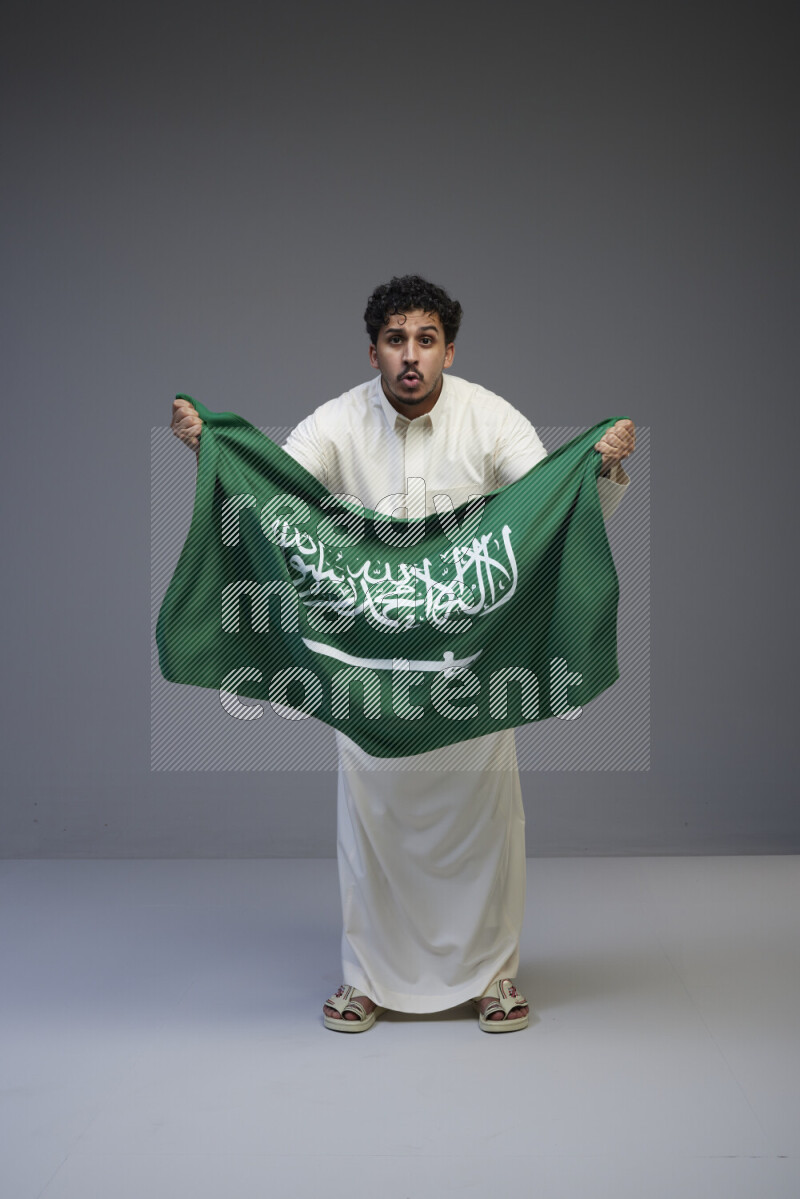 A Saudi man standing wearing thob holding big Saudi flag on gray background