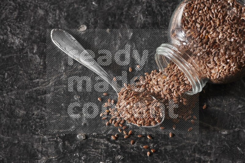 A glass spice jar full of flaxseeds flipped and seeds spread out with a metal spoon full of the seeds on a textured black flooring