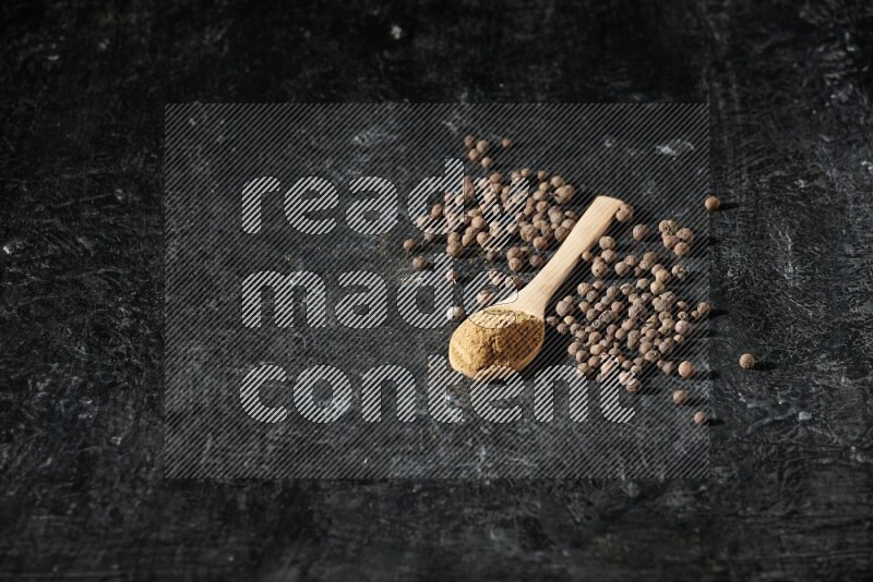 Wooden spoons full of all spice powder and allspice whole balls beside it on a textured black flooring