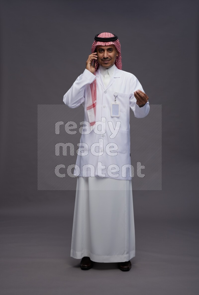 Saudi man wearing thob with lab coat and shomag with pocket employee badge standing talking on phone on gray background