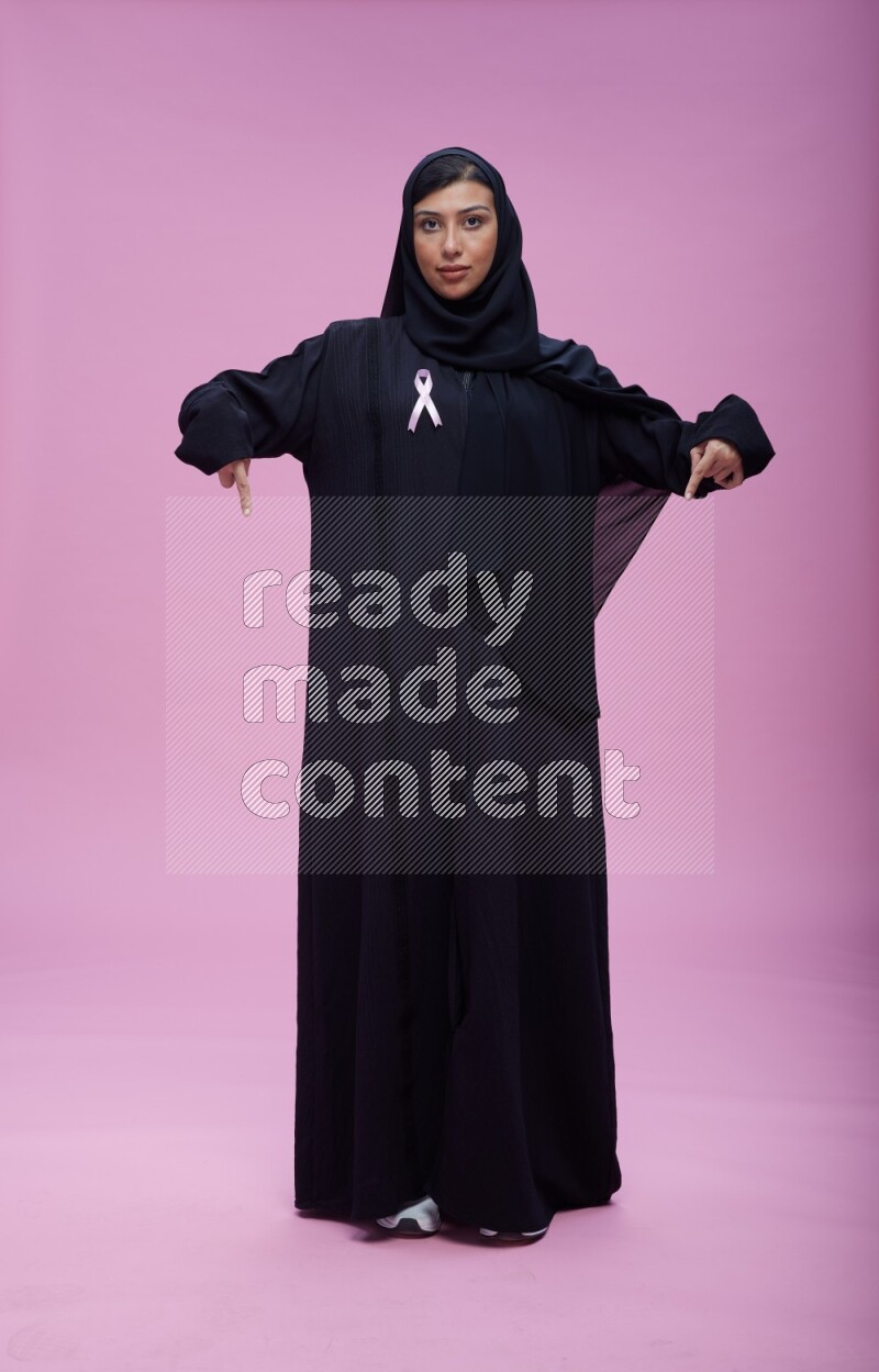 Saudi woman wearing pink ribbon on Abaya standing interacting with the camera on pink background