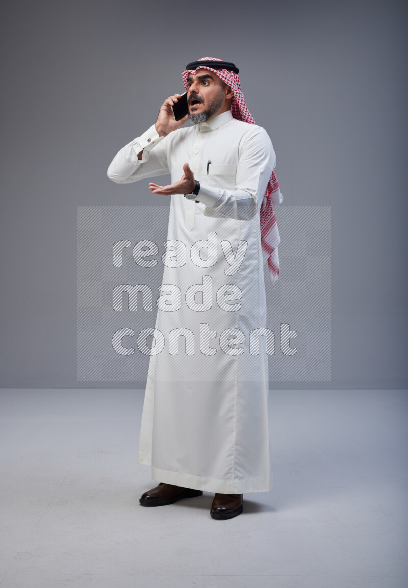 Saudi man Wearing Thob and red Shomag standing talking on phone on Gray background