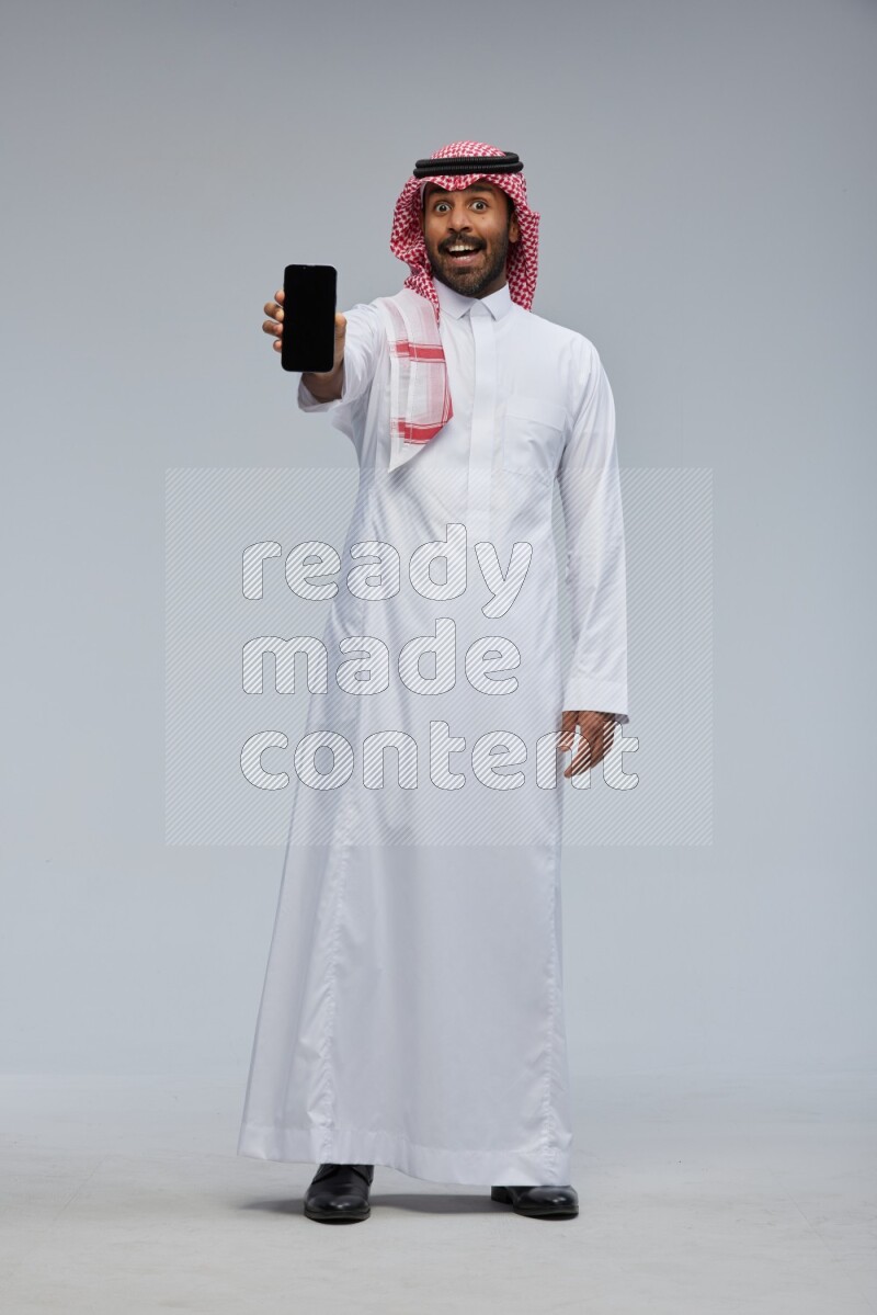 Saudi man Wearing Thob and shomag standing showing phone to camera on Gray background