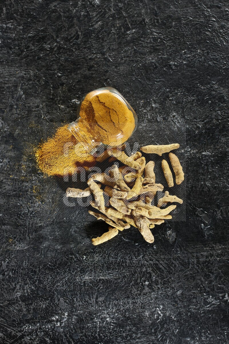 A flipped glass spice jar full of turmeric powder and powder spilled out of it with dried whole fingers on textured black flooring