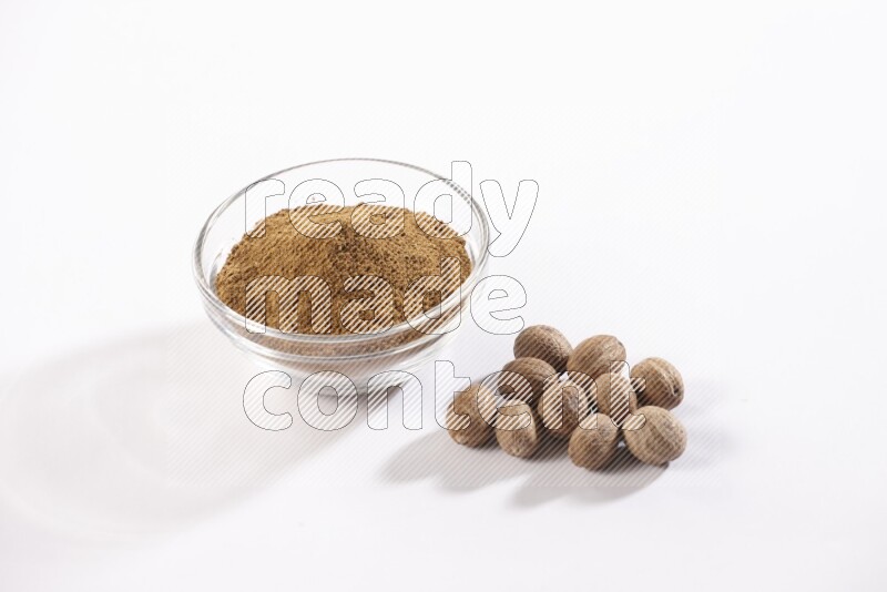 A glass bowl full nutmeg powder with the whole seeds beside it on a white flooring