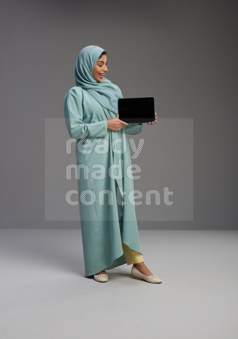 Saudi Woman wearing Abaya standing showing tablet to camera on Gray background